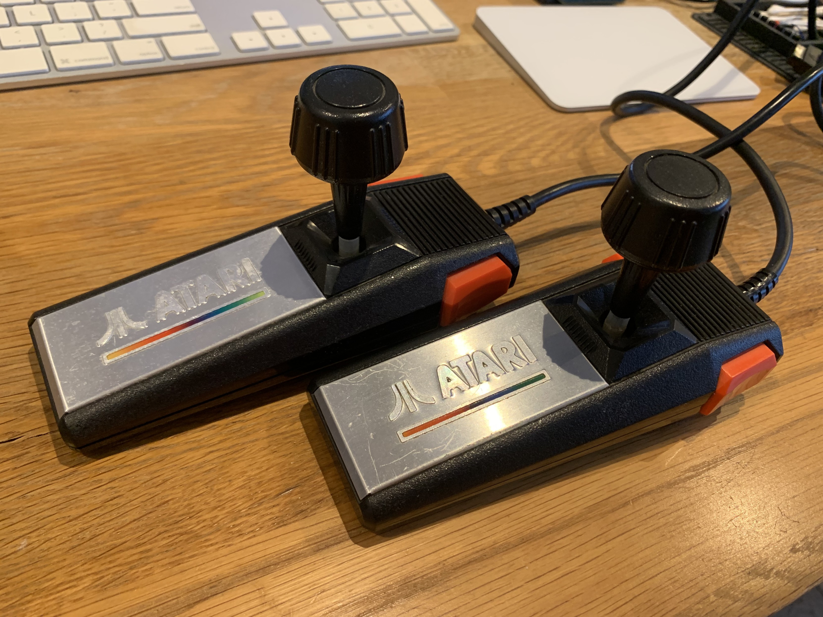 Two Atari joysticks, with a shiny metal plaque with a rainbow line, above it the Atari logo. The joysticks are to the right of the plaque. Two red buttons are at the side of the controller.