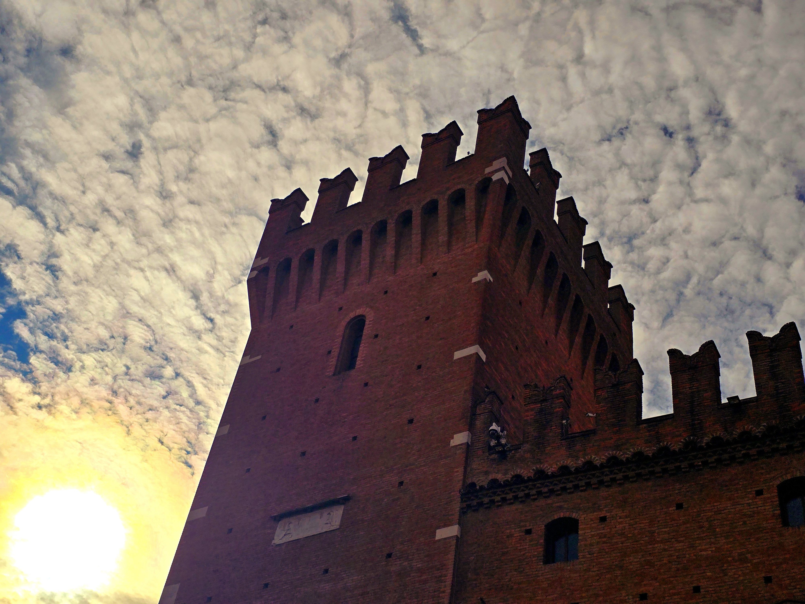 The silhouette of a medieval brick tower against a dramatic, cloudy sky during sunset in Ferrara, Italy. The crenellated top of the tower stands starkly against the textured clouds, illuminated by the warm, golden glow of the sun peeking from the lower left. The contrast between the ancient architecture and the vibrant sky creates a striking and timeless atmosphere, capturing the essence of Ferrara's historical charm at dusk.