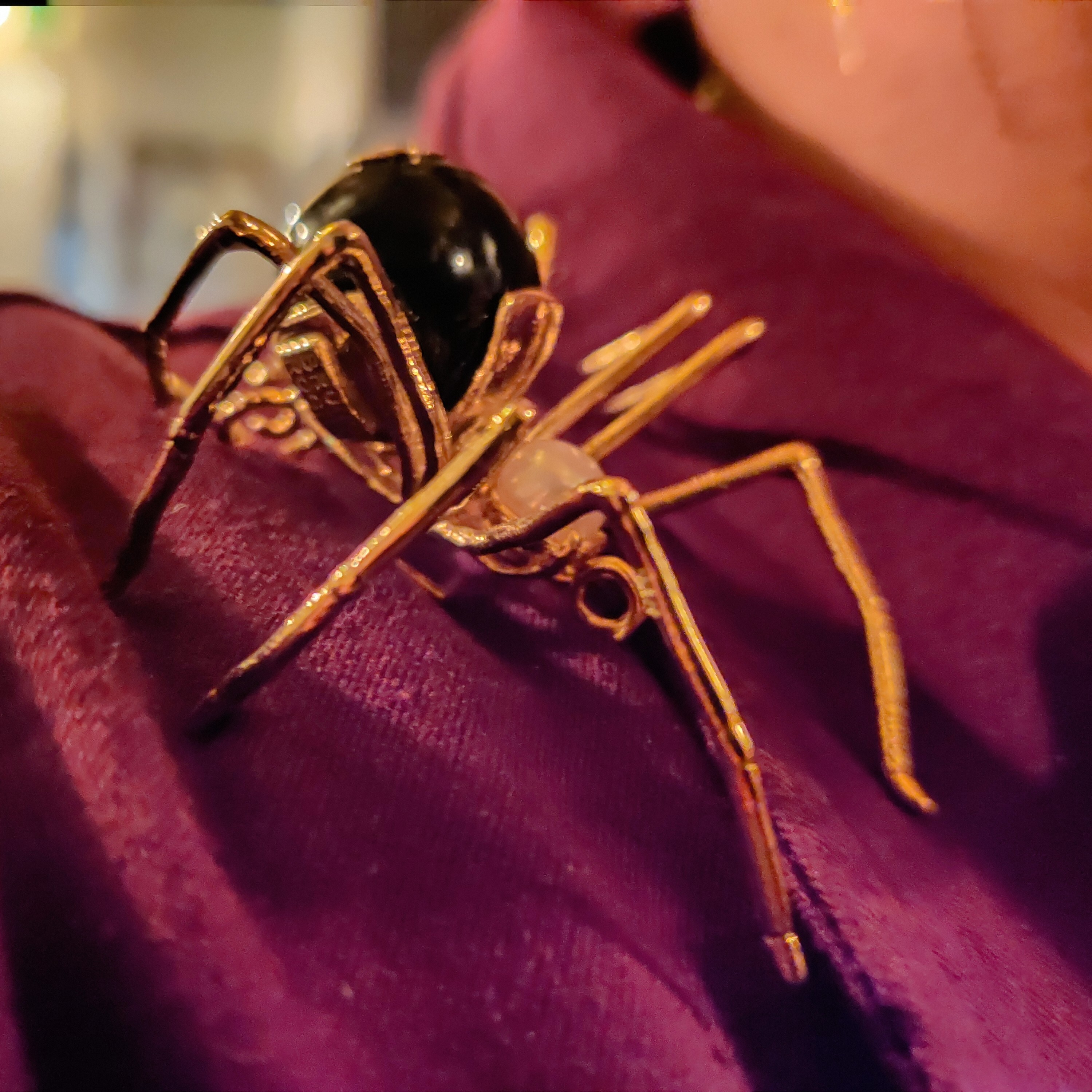 A golden spider brooch with a pearl head and a black onyx abdomen on maroon fabric.