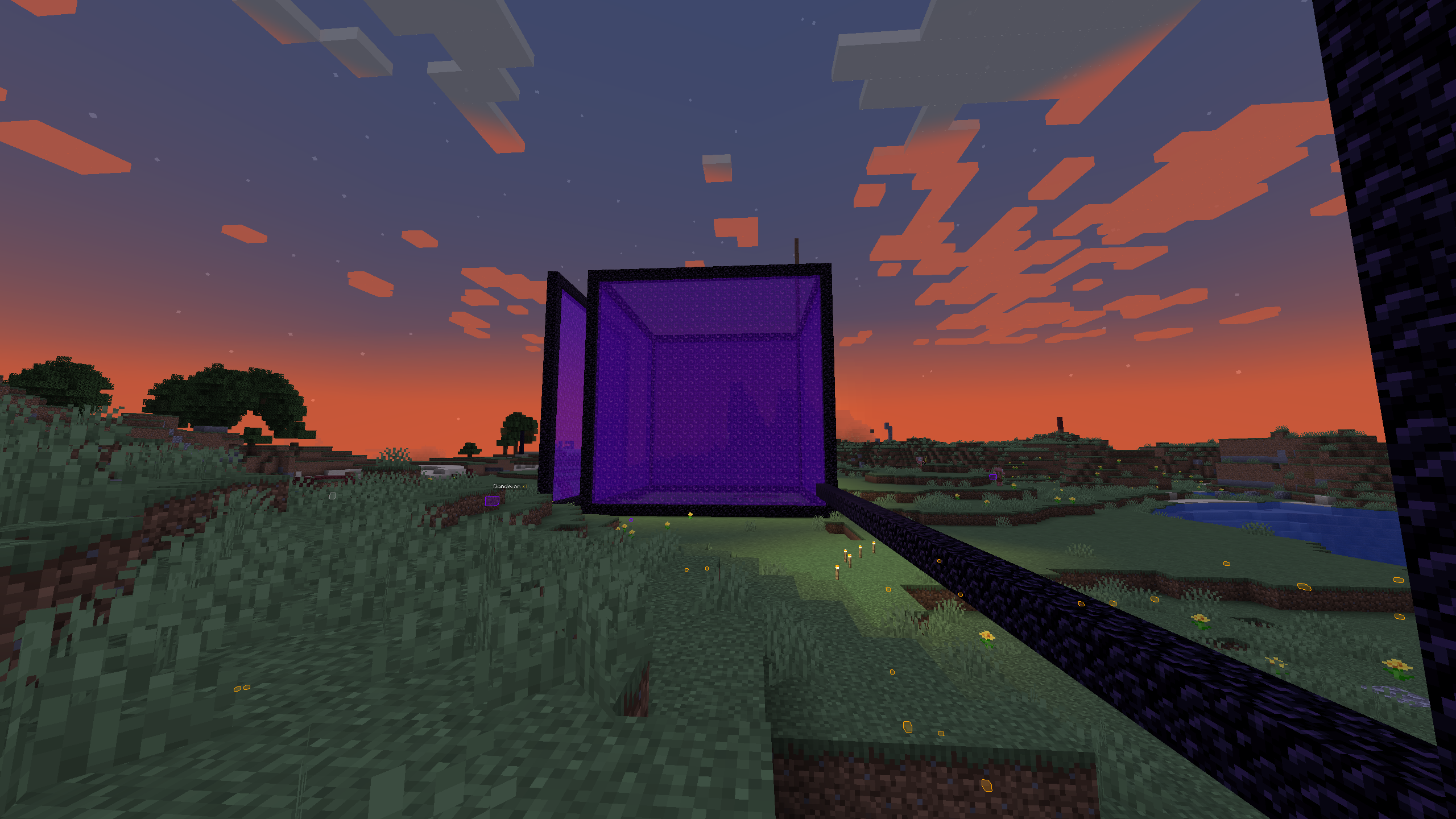 A screenshot from the game Minecraft. It shows a large, cube-shaped portal, likely a Nether portal, constructed with obsidian blocks and glowing with a purple interior. The structure appears much larger than a typical portal, indicating it was custom-built on a larger scale.

The setting is an open grassy landscape with a few hills, trees, and a small body of water in the distance. The sky is depicted at sunset, with a gradient from orange near the horizon to dark blue higher up, scattered with blocky clouds typical of Minecraft. There are a few scattered torches on the ground, illuminating parts of the scene.

The portal seems to be the focal point of the image, standing prominently against the vibrant sky as the day turns to night in the game world.