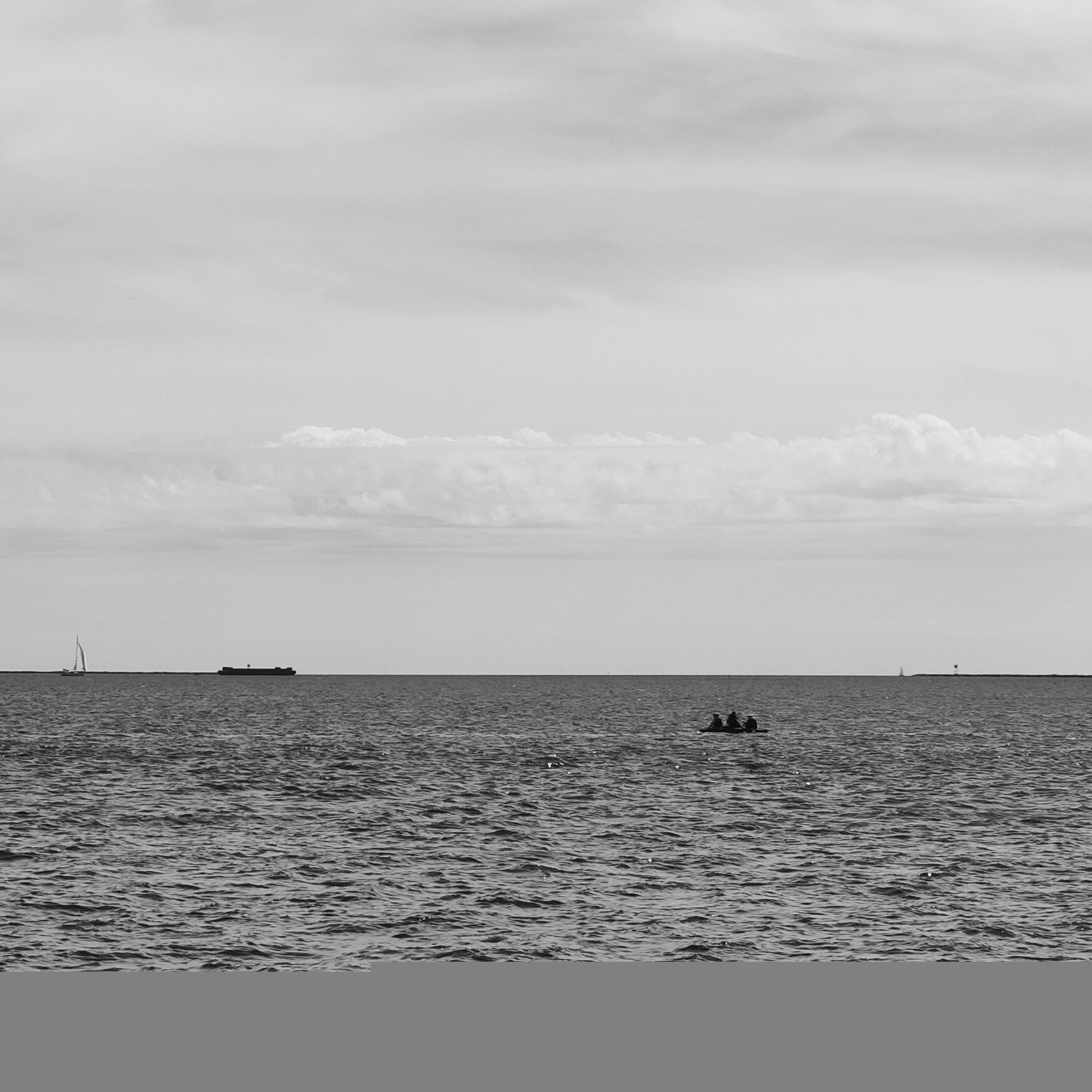 Black and white photo of New Haven Harbor from the West Haven store, showing a small boat with three fishermen in the middle ground and a barge and a sailboat on the horizon.