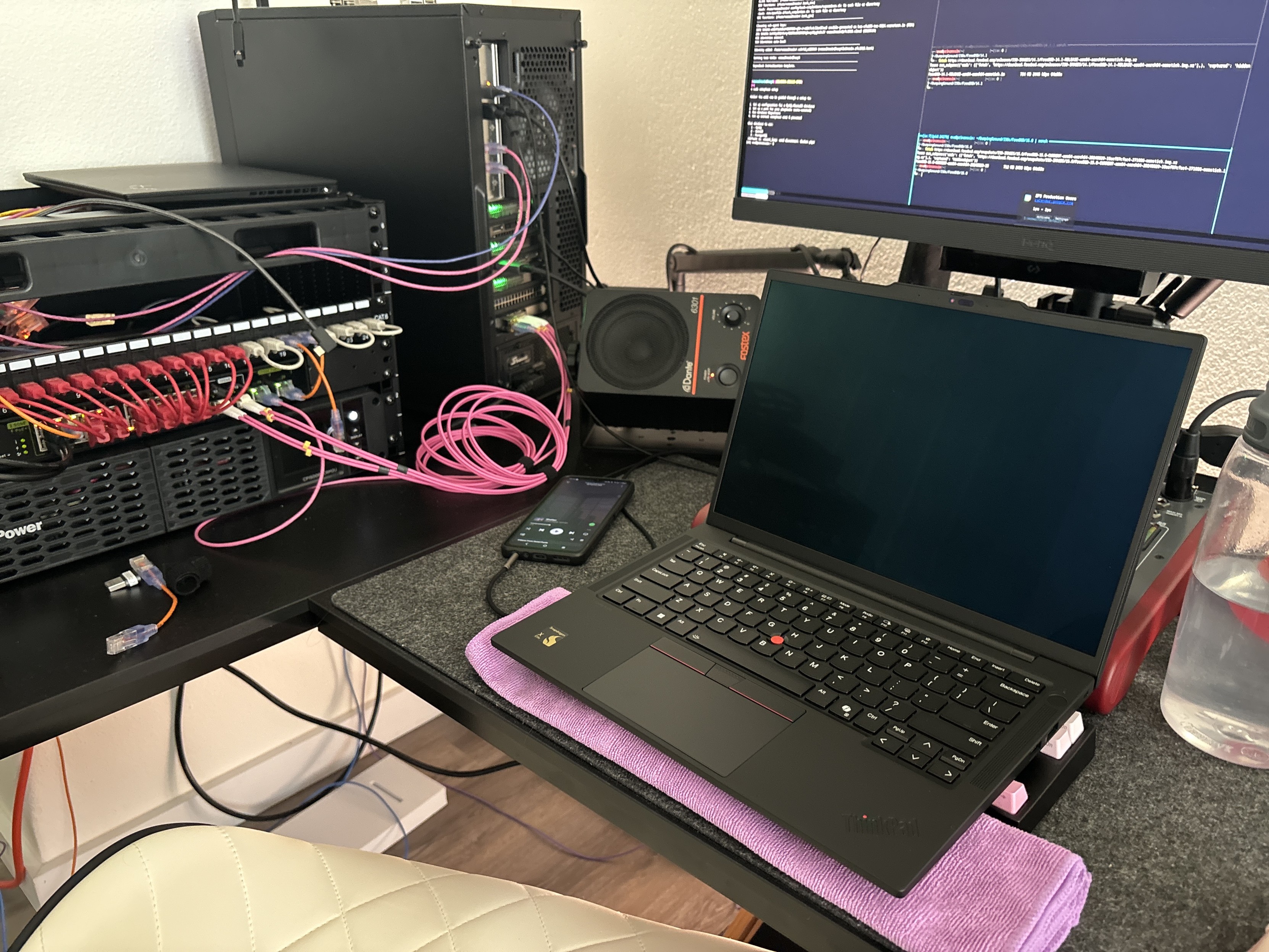 A workspace featuring a laptop with a dark screen, a server rack with numerous pink cables, an external monitor displaying code, and a smartphone. Additional items include a water bottle and a sound system.