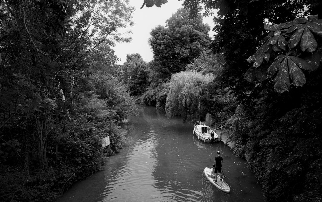 Black and white daylight photo.
People standing on a fragile board over the river deliberately engulfing themselves into the not so far wild unknown.